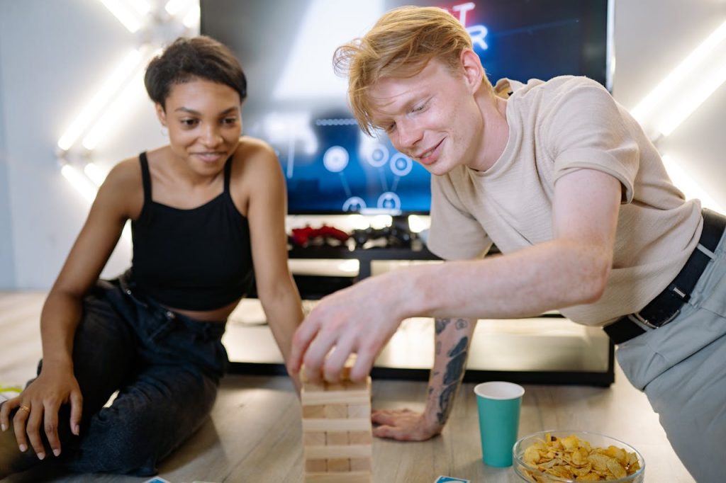friends-playing-jenga-9068978 Two friends playing Jenga and enjoying snacks indoors, creating a fun and relaxed atmosphere.